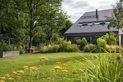 ohľad od pergoly s grilom cez viacúčelovú trávnatú plochu s kovovým ohniskom (vpravo) smerom k domu. Za nízkou bariérou z kríkov a trvaliek sa ukrýva terasa pri dome. Vľavo kmeň stromu obklopuje originálna guľatá drevená lavica, ktorá ponúka jedno z mnohých miest na posedenie na záhrade